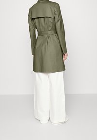 Osoba ubrana w oliwkowy płaszcz trench z paskiem i szerokie białe spodnie, stojąca tyłem do kamery na jednolitym tle.