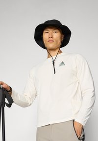 Langärmliges cremefarbenes Hemd mit einem kleinen grünen Berg-Logo, kombiniert mit einem schwarzen Bucket Hat. Das Modell hält einen Trekkingstock.