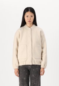 Veste bomber en polaire ivoire avec un col rond, deux poches avant, un ourlet élastiqué et des poignets côtelés, présentant une surface douce et texturée.