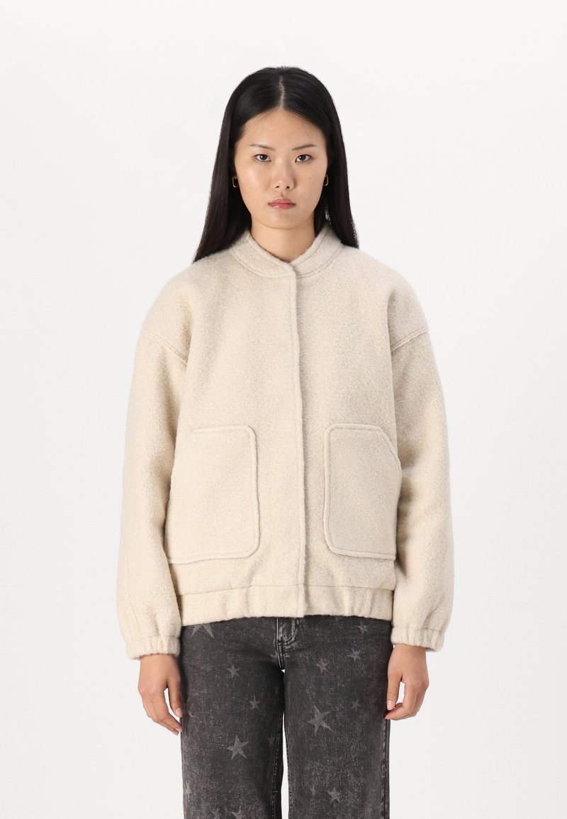 Veste bomber en polaire ivoire avec un col rond, deux poches avant, un ourlet élastiqué et des poignets côtelés, présentant une surface douce et texturée.