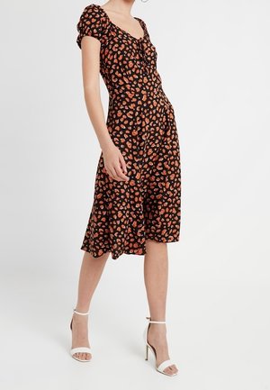 Nainen pukeutuneena mustaan midi-mekkoon, jossa on oranssinvärinen leopardikuvio, puhvimaiset hihat ja valkoiset nilkkaremmi-korkokengät, seisoo valkoisella lattialla.