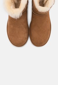 Brązowe botki zamszowe z miękką, kremową podszewką z owczej skóry, zaokrąglonym noskiem i elastyczną gumową podeszwą. Bez widocznych zapięć.