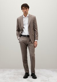 Beige Anzug mit subtiler Textur, weißem Hemd, schwarzem Gürtel und glänzenden schwarzen Dressschuhen. Der Anzug hat eine taillierte Passform und Schlagklappen.