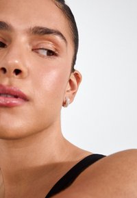 Sudraba riņķveida auskars ar mirdzošiem kristāla akcentiem. Modelis ir ar gludu, starojošu ādu un valkā melnu topu ar siksniņām.