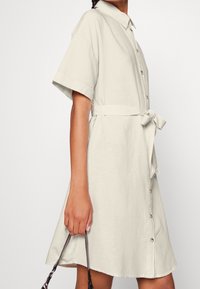 Abito camicia beige chiaro realizzato in tessuto testurizzato, caratterizzato da maniche corte, una cintura con fiocco in vita e una gonna svasata.