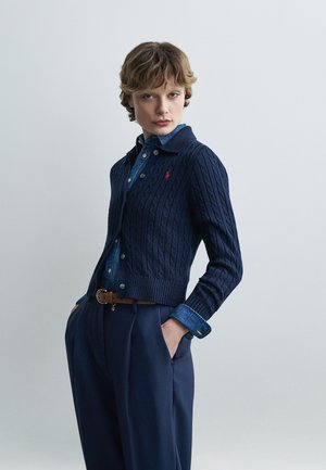 Ung person med kort hår iført en marineblå kabelstrikket cardigan over en blå skjorte med knapper og marineblå bukser, hænderne i lommerne, neutral baggrund.