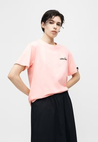 Νεαρός με κοντά σκούρα βρεγμένα μαλλιά, φορώντας ανοιχτό ροζ μπλουζάκι ellesse και μαύρο παντελόνι, στέκεται με τα χέρια στη μέση σε λευκό φόντο.