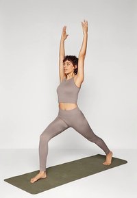 Világosbarna sportos szett, amely egy ujjatlan felsőből és magas derekú leggingsből áll, egy személy viseli jógapózban egy olívazöld matracon.