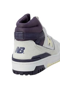 New Balance sneaker met een paarse textielkraag, grijze suède bovenkant, witte tussenzool en zwarte rubberen loopzool met textuur en logo-detail.
