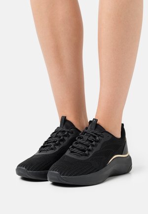 Fødder iført sorte mesh-sneakers med mønstrede snørebånd og en tyk sål, stående på en hvid overflade.