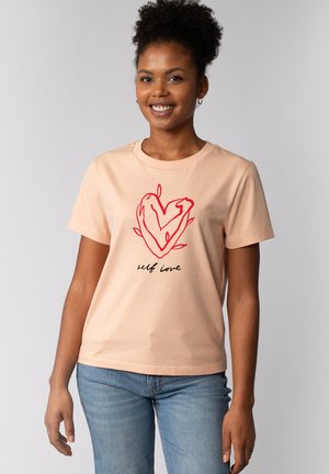 T-shirt à manches courtes de couleur pêche avec un design de cœur rouge et le texte "self love" en noir. Tissu doux, col rond, coupe régulière.