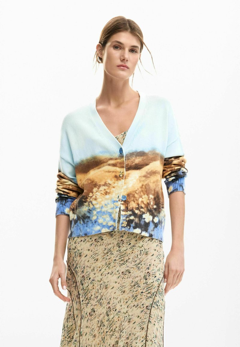 Világoskék kötött kardigán tájképes mintával, V-nyakkal, gombolással és bordázott mandzsettával; virágmintás ruha fölött viselve.