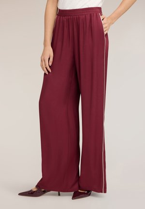 Femme portant un pantalon bordeaux à jambes larges avec taille élastique et bande latérale, associé à des talons pointus bordeaux et un haut blanc.
