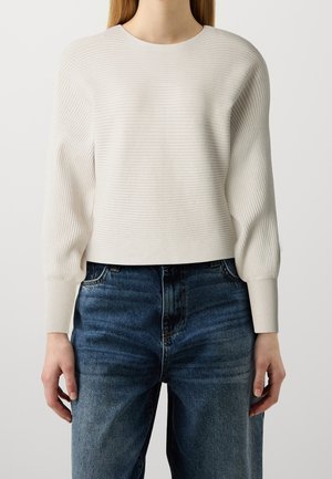 Femme portant un pull côtelé crème à manches longues bouffantes et un jean bleu foncé taille haute sur un fond uni.