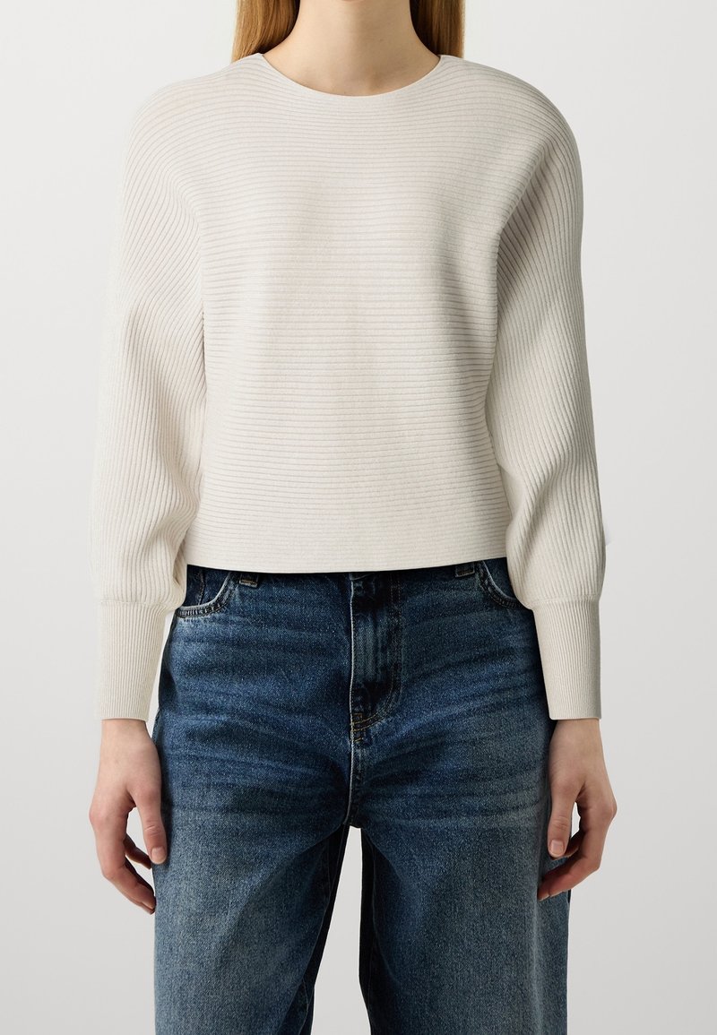 Femme portant un pull côtelé crème à manches longues bouffantes et un jean bleu foncé taille haute sur un fond uni.
