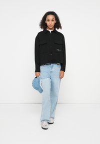Black cropped jacket with two chest pockets, button closure, and embroidered logo. Paired with light blue wide-leg jeans and a blue cap.
