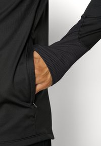 Schwarzes Langarmshirt mit Seitentasche und gerippten Bündchen. Weiches Material, nahtloses Design und strukturierte Nähte entlang der Kanten.