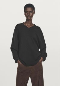 Maglione nero in maglia oversize con scollo a V, caratterizzato da maniche ampie, indossato con pantaloni in suede marrone. Tessuto semplice e strutturato.