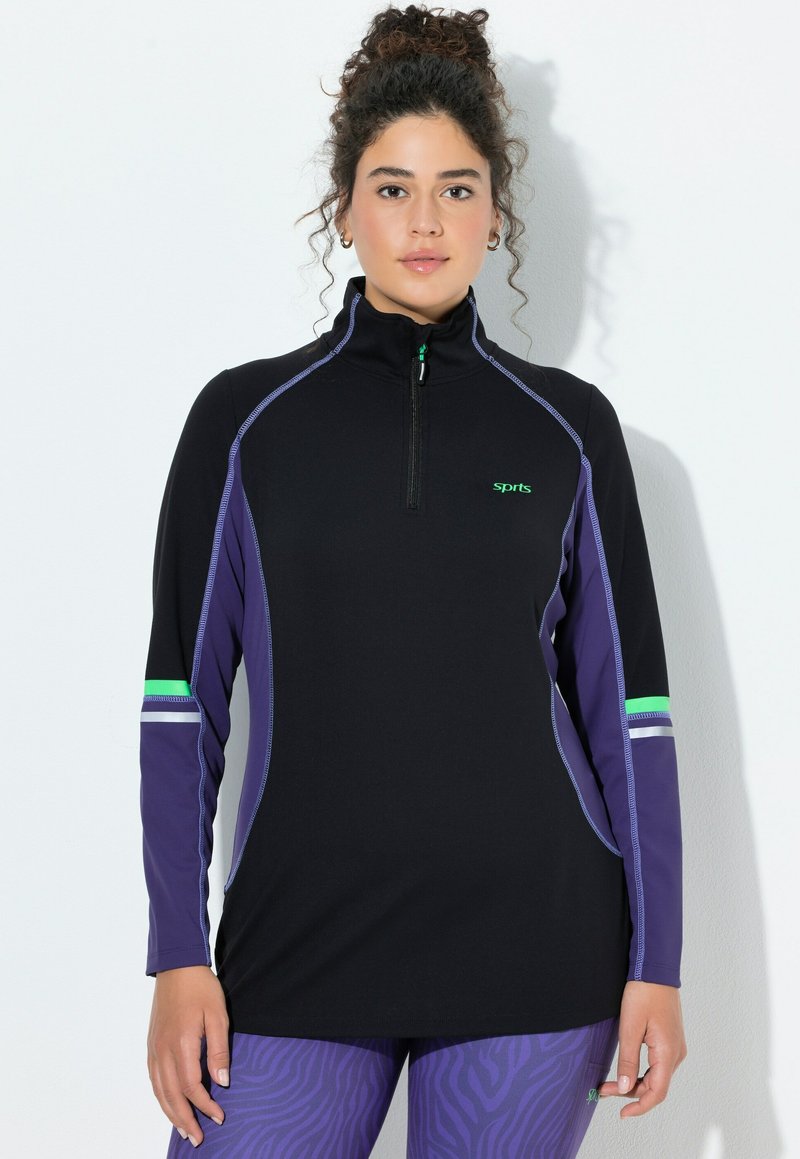 Žena s kudrnatými vlasy v černém a fialovém sportovním tričku s rozepínacím límcem a fialových legínách s vzorem, stojící před bílou zdí.