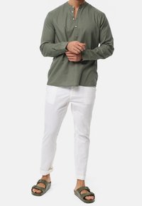 Camisa de manga larga verde oliva con cuello mandarín y tapeta de botones, combinada con pantalones blancos ajustados y sandalias tipo slip verdes oliva.