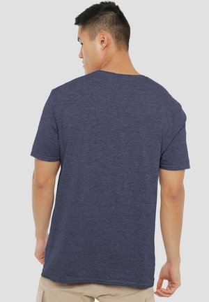 Young man facing away wearing a plain dark blue short-sleeve t-shirt and beige pants against a light background.