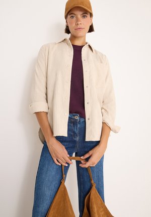 Camicia in corduroy beige con chiusura a bottoni, indossata sopra un maglione bordeaux. Abbinata a jeans blu e una borsa in camoscio marrone con manici.