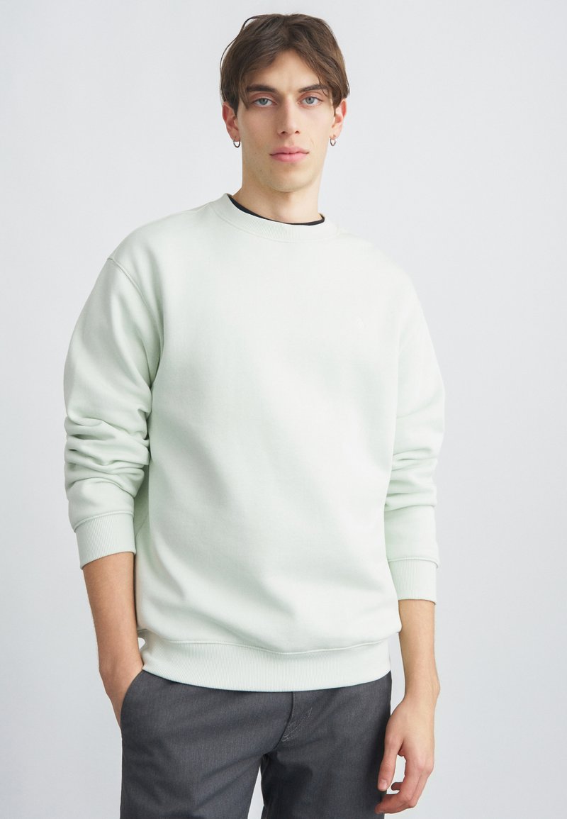 Sweat-shirt vert menthe clair à col rond, fabriqué en tissu lisse avec poignets et ourlet côtelés. Porté par-dessus une chemise noire, associé à un pantalon foncé.