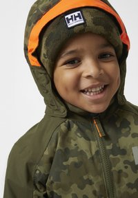 Chaqueta de camuflaje verde con ribete naranja y cierre de cremallera. La chaqueta tiene capucha y un parche con logo en la parte delantera. Gorro de punto de camuflaje.