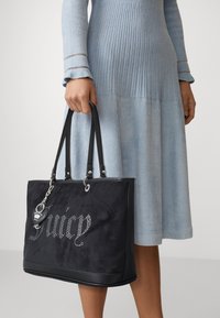 Woman in a light blue ribbed dress holding a black tote bag with "Juicy" in rhinestones and a heart-shaped keychain charm.