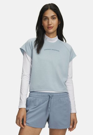 Vrouw draagt een lichtblauw Under Armour mouwloos topje over een wit shirt met lange mouwen en bijpassende lichtblauwe short met trekkoord, staand tegen een witte achtergrond.