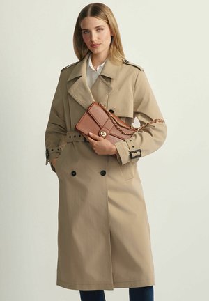 Kobieta w beżowym płaszczu trencz, trzymająca brązową skórzaną torebkę ze złotym łańcuszkiem na tle jasnego, jednolitego tła.