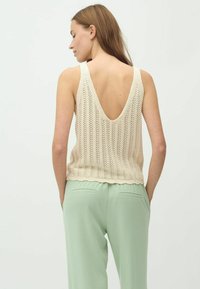 Beiger gestrickter Tank-Top mit tiefem Rückenausschnitt, ausgestattet mit einem strukturierten Muster, kombiniert mit hellgrünen schmal geschnittenen Hosen mit Gesäßtaschen.