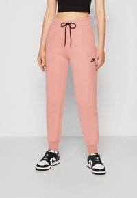 Růžové bavlněné tepláky s elastickým pasem, černým stahovací šňůrou, bočními kapsami a malým černým logem Nike na horní části stehna.