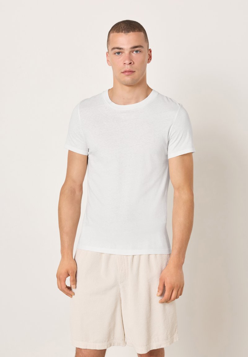 Maglietta bianca in cotone con scollo rotondo e maniche corte, abbinata a pantaloncini beige chiaro con trama. Design semplice e aderente.