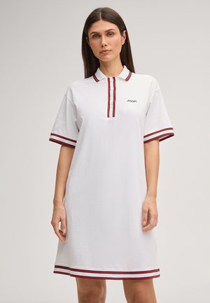 Kobieta w białej sukience polo z krótkim rękawem z czerwono-czarnymi paskami na kołnierzyku, rękawach, plisie z guzikami i na dole sukienki.