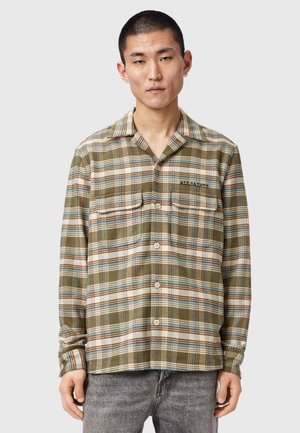 Mężczyzna w oliwkowo-zielonej i beżowej kratkowanej koszuli na guziki z kieszeniami na piersi oraz w szarych dżinsach, stojący na jednolitym tle.