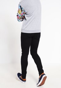 Sudadera gris con estampado colorido en las mangas, pantalones ajustados negros y zapatillas azul marino tipo slip-on con detalles en naranja, visto desde la parte trasera.