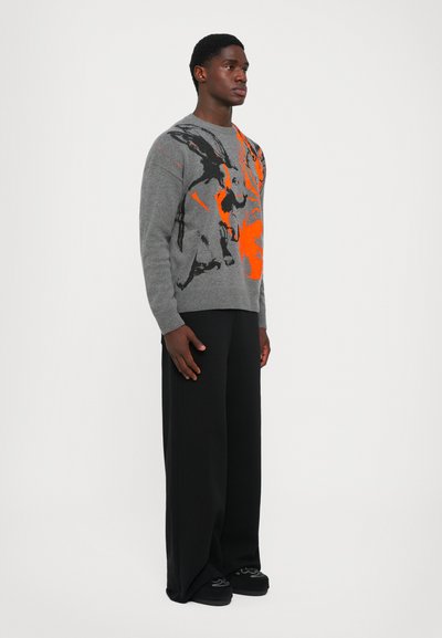 Grauer Pullover mit abstraktem orange-schwarzem Design, runder Ausschnitt, strukturierter Stoff. Kombiniert mit schwarzen weiten Hosen und schwarzen Schuhen.
