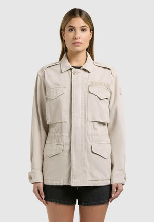 Femeie în picioare cu fața spre înainte, purtând o jachetă bej cu patru buzunare cu clapetă și pantaloni scurți negri, pe un fundal simplu, deschis la culoare.