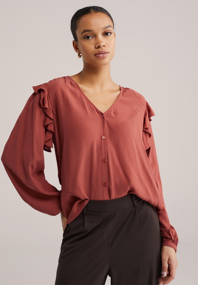 Blouse couleur rouille avec détails à volants sur les manches, col en V et fermeture à boutons. Tissu léger avec une texture douce. Associée à un pantalon foncé.