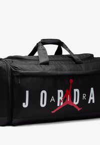 Sac de sport noir avec un dessus zippé, arborant le logo rouge "Jumpman" et le texte "JORDAN" en blanc. Tissu doux, poignées robustes pour un transport facile.