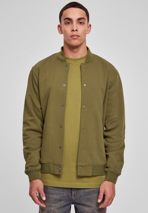Young man wearing an olive green snap-button jacket over a matching t-shirt and blue jeans, standing against a plain light gray background.