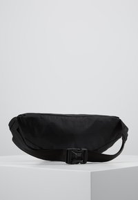 Sac en bandoulière en tissu noir avec une texture lisse, de forme rectangulaire, une sangle réglable et une fermeture à boucle latérale.