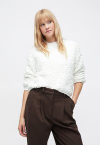 Valkoinen pörröinen neule, jossa on teksturoitu pinta, pyöreä pääntie ja lasketut olkapäät, yhdistettynä ruskeisiin korkeavyötäröisiin housuihin.