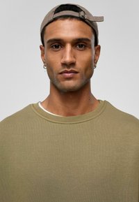 Olivengroene crewneck sweatshirt gemaakt van gestructureerde stof, gelaagd over een wit shirt, gedragen met een beige pet. Het model heeft kort haar en een oorbel.