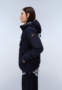 Frau im Profil trägt eine dunkelblaue Kapuzenjacke mit der Hand in der Tasche, darüber ein kariertes Hemd, vor einem schlichten hellen Hintergrund.
