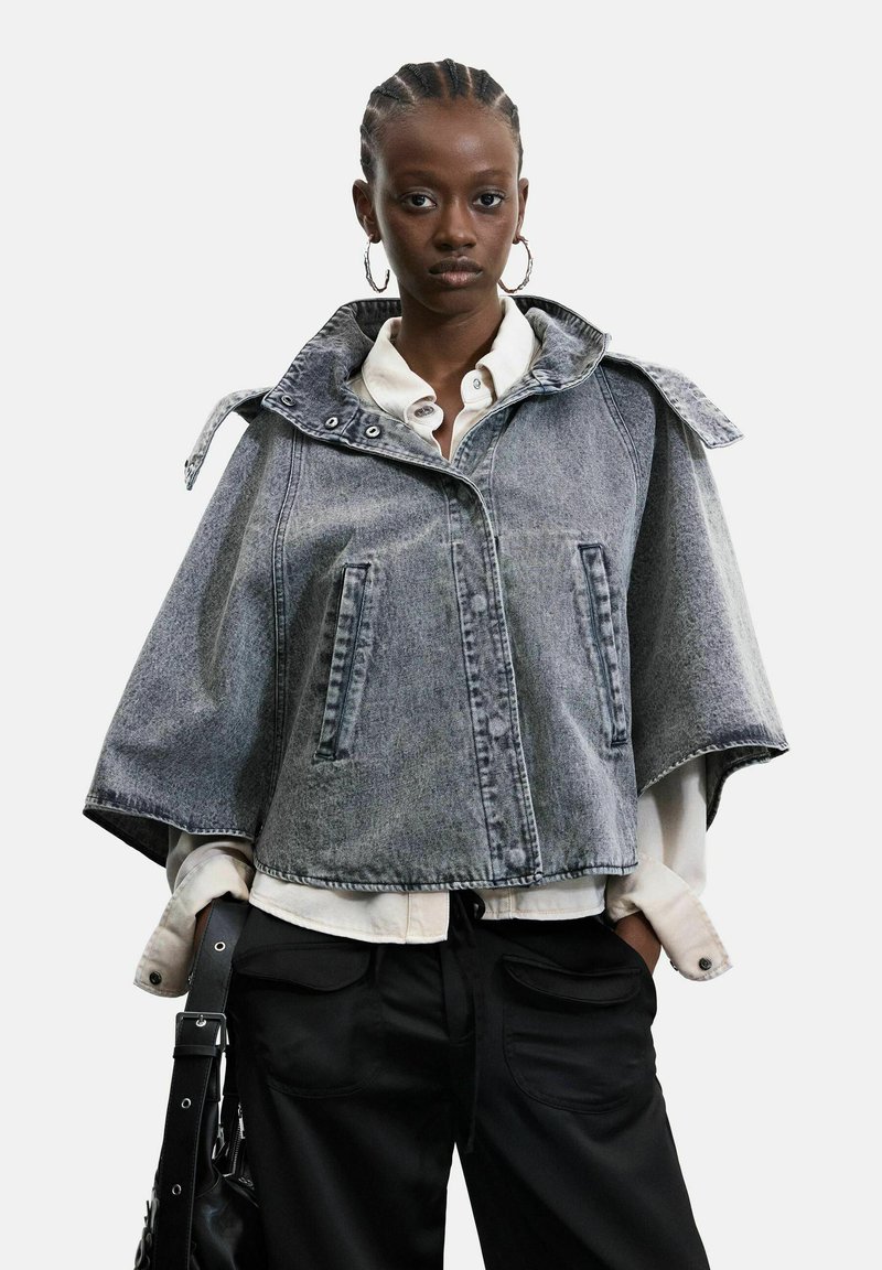Femme aux cheveux tressés porte une cape en denim gris, une chemise blanche, un pantalon noir, de grandes boucles d'oreilles et tient un sac noir avec une sangle à boucle.