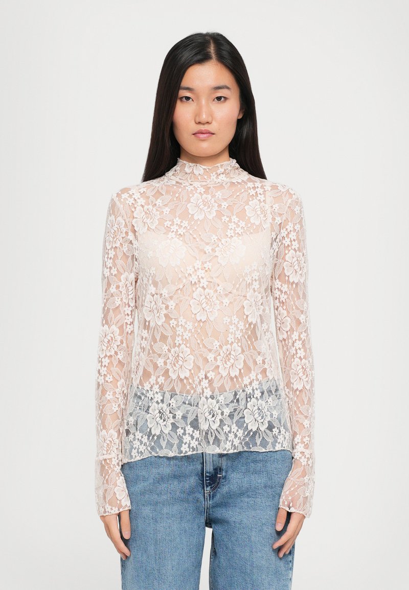 Beige en witte kanten lange mouw top met bloemmotief, hoge hals en aansluitende manchetten, gedragen over een lichtblauwe denim onderkant.
