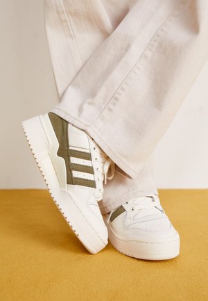 Osoba nosząca beżowe spodnie i białe trampki z oliwkowozielonymi paskami, stojąca na musztardowożółtej wykładzinie.