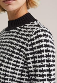 Stickad tröja med en svart ribbad krage; har ett geometriskt mönster i svart och vitt, med växelvis strukturerade rader.